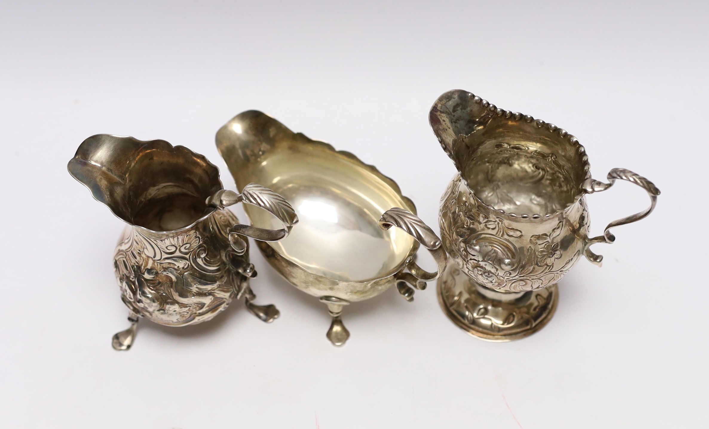 A late George II silver sauceboat with flying scroll handled, London, 1759 and two Georgian silver cream jugs, with later embossed decoration (a.f.)